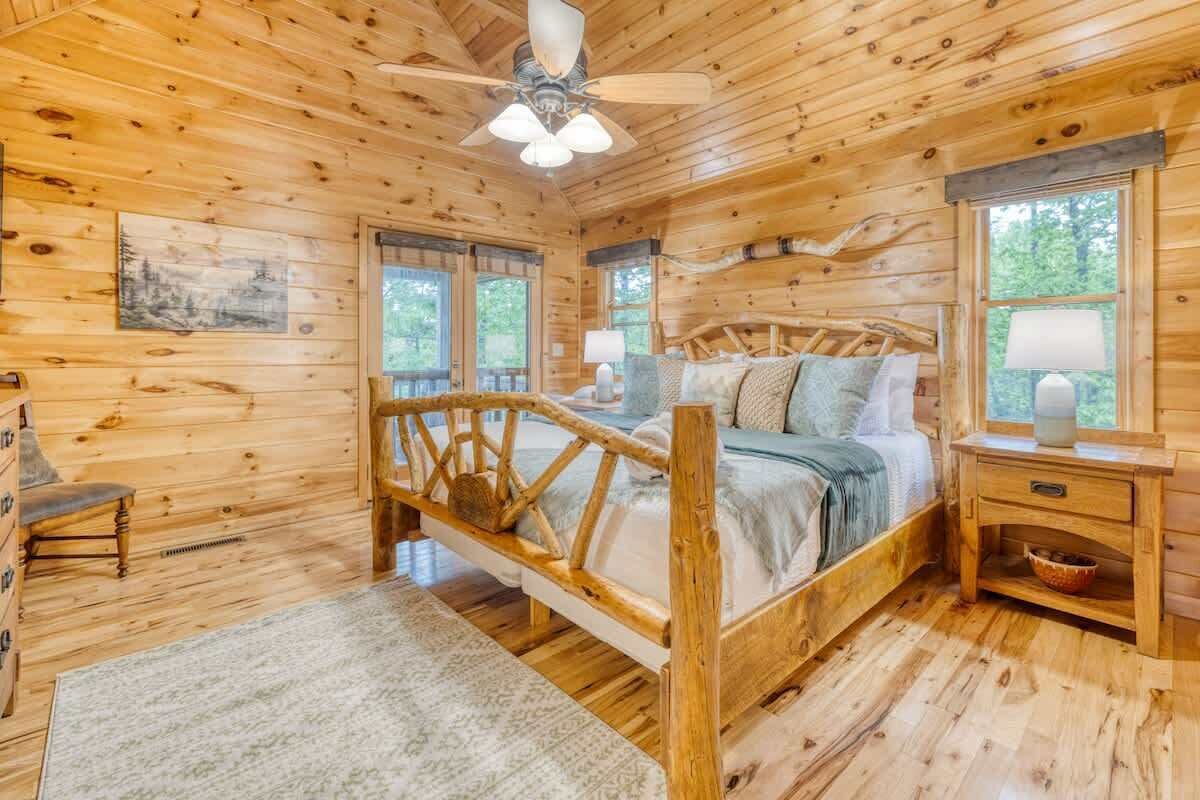 A wooden cabin bedroom with a bed, a nightstand, a chair, a ceiling fan, and windows.