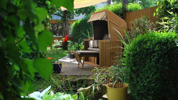 A wooden deck with a wicker chair and a small table, surrounded by lush greenery and a wooden fence. - Holiday Apartment Langkamp (Neuenkirchen)