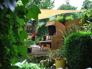 A wooden deck with a wicker chair and a small table, surrounded by lush greenery and a wooden fence. - Holiday Apartment Langkamp (Neuenkirchen)