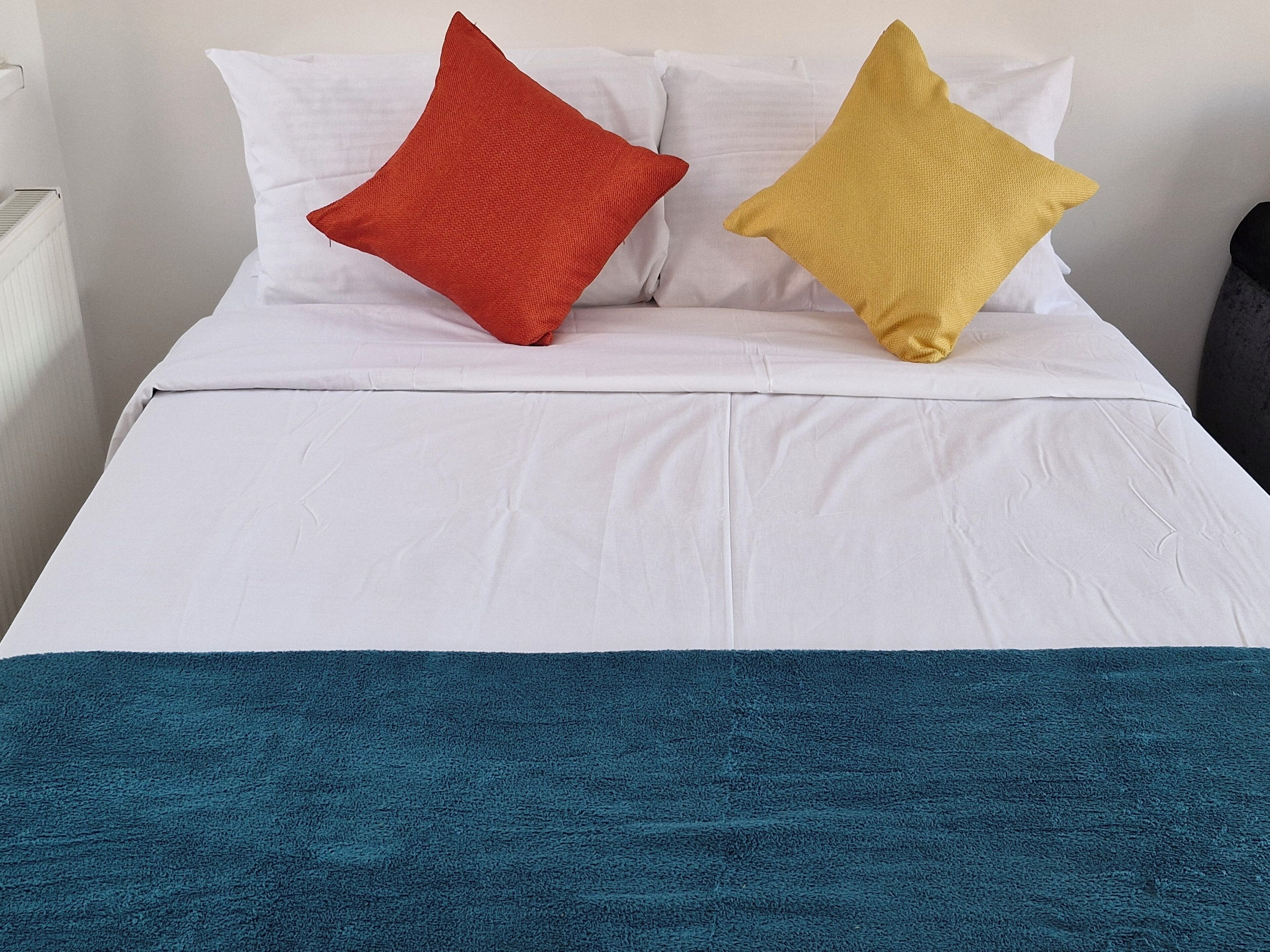 A bed with white bedding, a teal throw blanket, and two pillows, one red and one yellow.