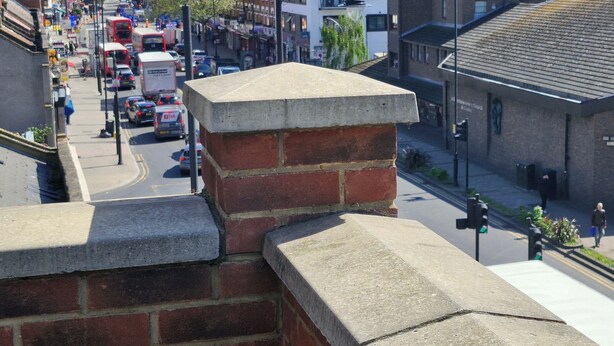 Une vue de rue avec un mur de briques et un bâtiment surmonté d’une tour horloge en arrière-plan.