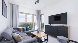 Un salon moderne avec un canapé gris, une petite table en bois, un pouf gris, un meuble TV noir et une grande fenêtre avec des rideaux.