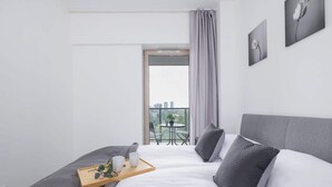 Une chambre d’hôtel moderne avec un grand lit, une literie grise, un plateau en bois agrémenté d’une plante, et une vue sur la ville depuis un balcon.