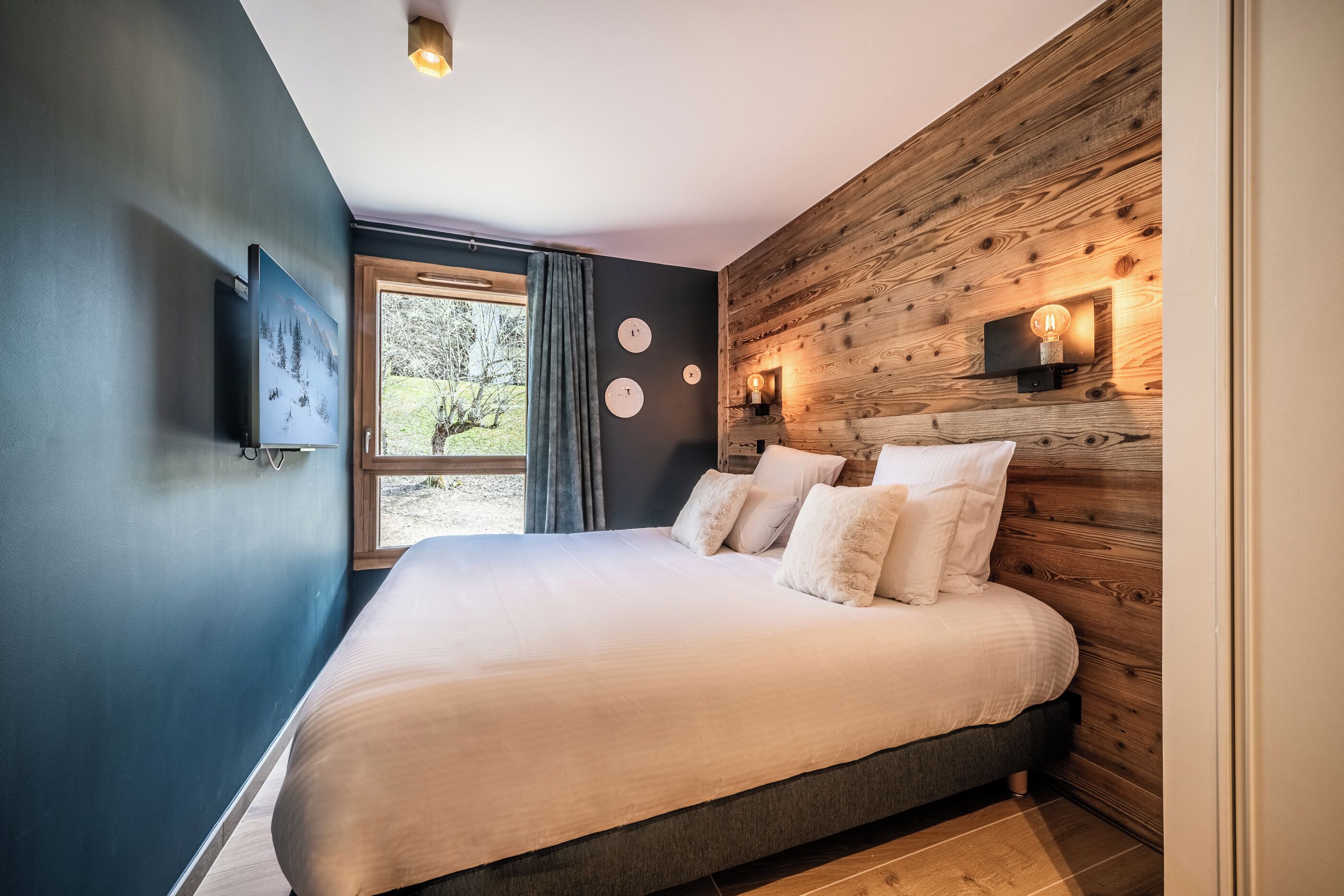 Een slaapkamer met een groot bed, een houten hoofdbord, een aan de muur bevestigde televisie en een raam met gordijnen.