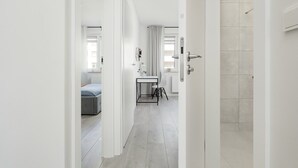 A modern hotel room with a bed, a small table with a chair, a bathroom with a shower, and a view of the street.