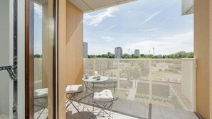 A balcony with a view of buildings and greenery, a small table with two chairs, and a white towel on the floor.