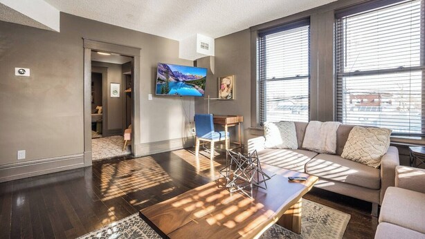 A spacious living room with a large window, a flat-screen TV, a wooden coffee table, and a beige sofa.