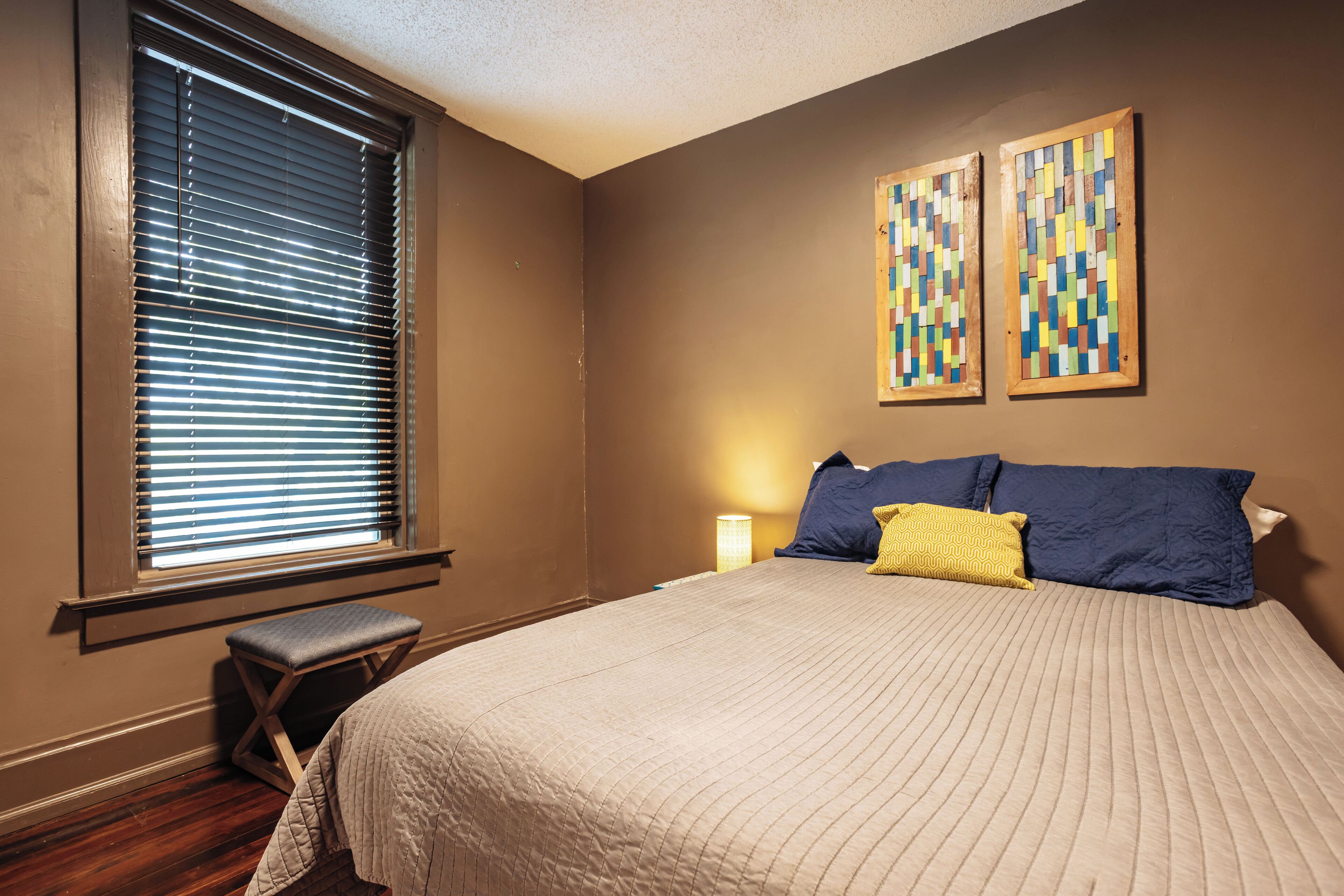 A bedroom with a bed, a window with blinds, a small stool, and two wall-mounted paintings.