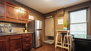 Una cocina con armarios de madera, un refrigerador de acero inoxidable, una pequeña mesa de madera con taburetes y una ventana con persianas.