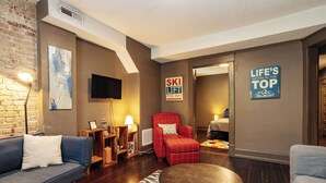 Una sala de estar moderna con un sofá, un sillón y una mesa de centro. Hay un televisor montado en la pared, un letrero que dice "SKI LIFT THIS WAY" y otro que dice "LIFE'S BETTER AT THE TOP".