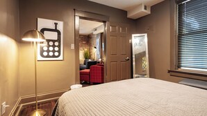 Un dormitorio con una cama grande, una lámpara de pie, una puerta que da a un baño y una ventana con persianas.