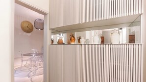 Een modern interieur met een glazen tafel en stoelen, een witte muur met verticale lijnen, en een plank met diverse keramieken vazen.