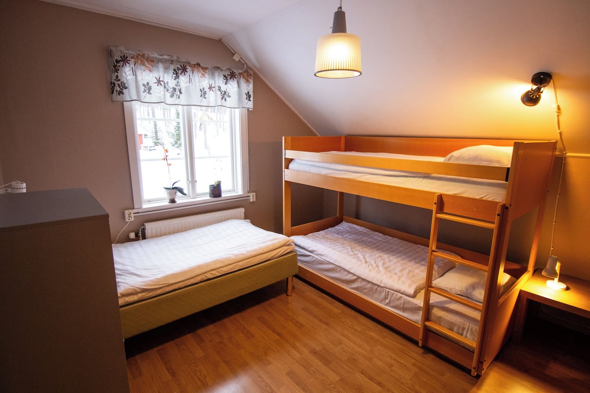 A dormitory room with bunk beds, a window with a curtain, a radiator, and a desk with a lamp.