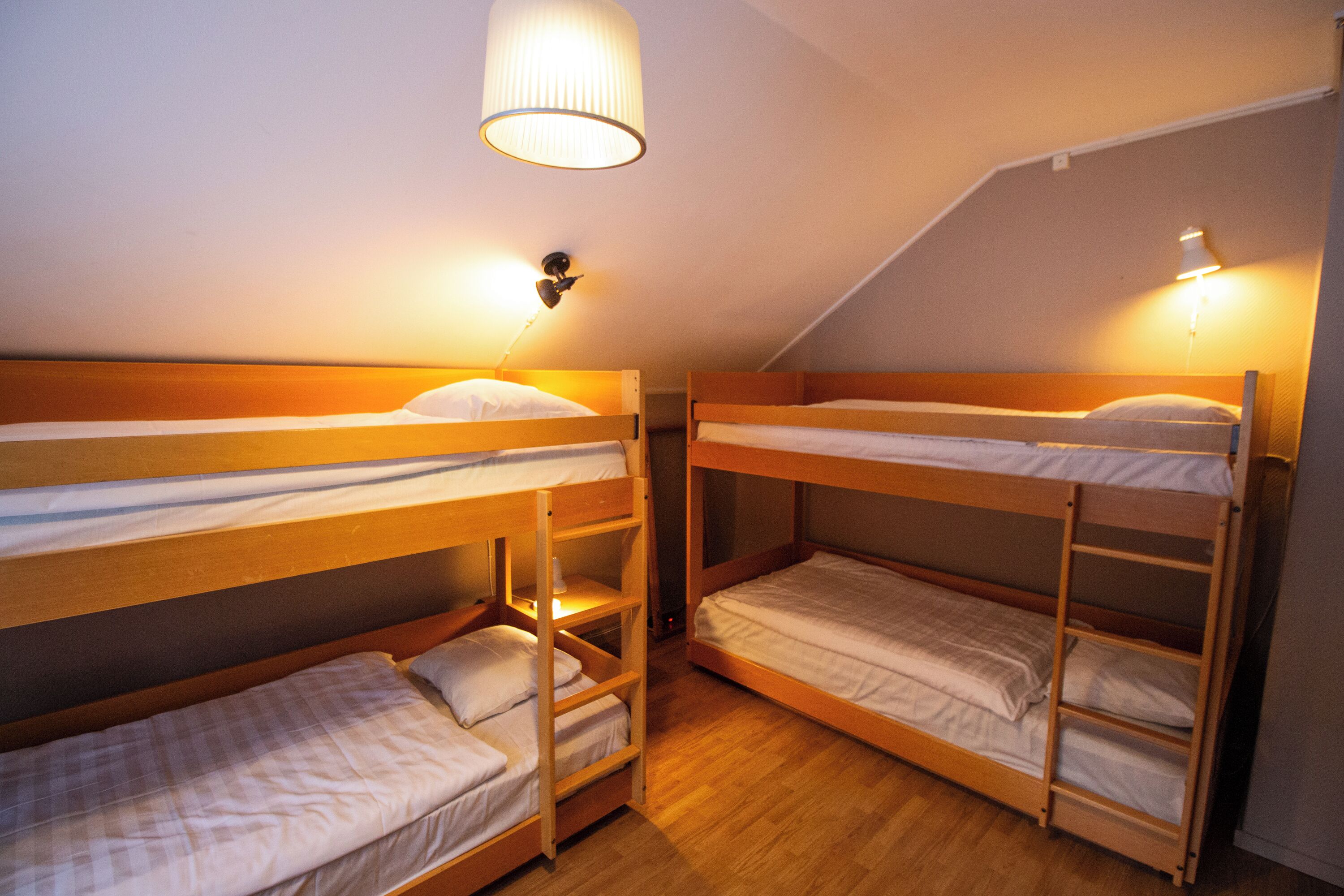 A dormitory room with two bunk beds, each with a shelf below. There is a ceiling lamp and a wall-mounted light fixture.