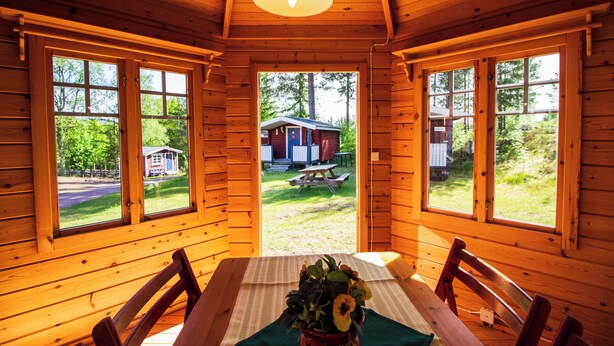 A wooden cabin with a dining table and chairs, large windows, and a view of a house and picnic table outside. - Gopshusgården - Rum och Stugor (Mora)