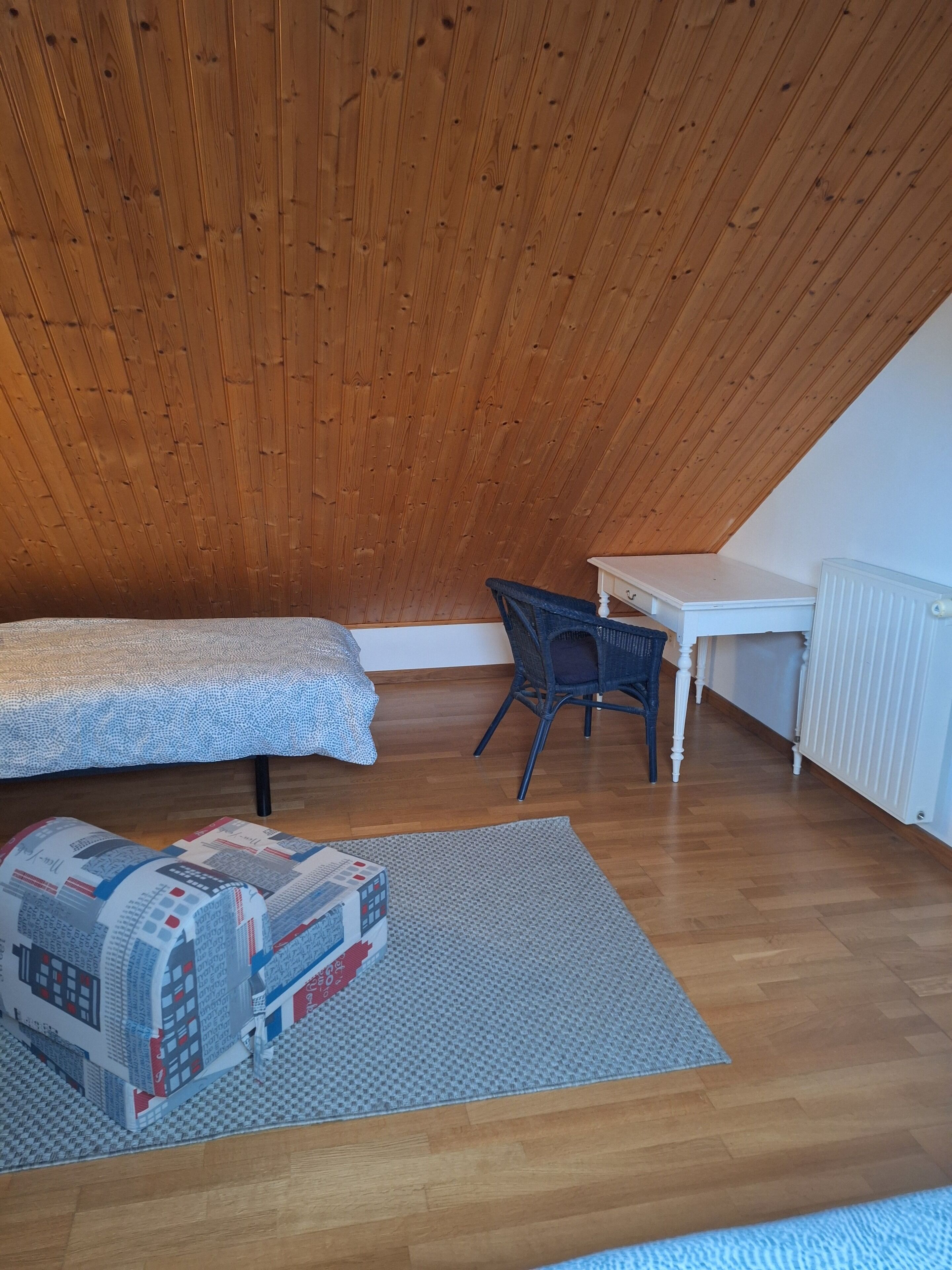 Una habitación con techo inclinado, una cama, un escritorio, una silla y un tapete estampado.