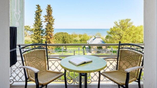A balcony with two wicker chairs and a small round table, overlooking a scenic view of the sea and a garden. - Strandvillen Wald & See (Zinnowitz)