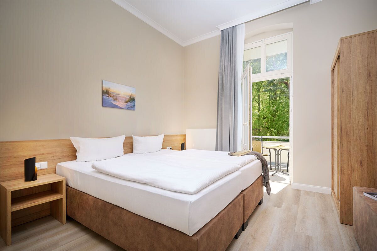 Ein Hotelzimmer mit einem großen Bett, einem Nachttisch, einem Fenster mit Vorhängen und Blick auf Grün.