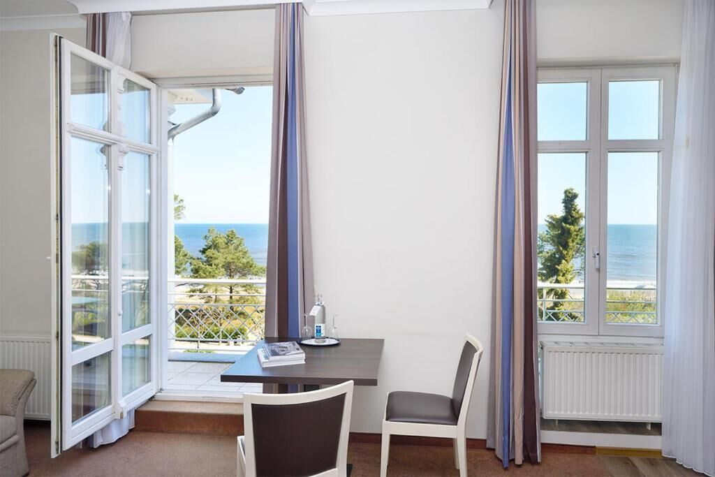 A room with a view of the sea, a table with a chair, and a window with curtains.