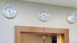 Ein Empfangsschalter mit drei Uhren an der Wand, eine sitzende Person dahinter und eine kleine, hängende Dekoration.
