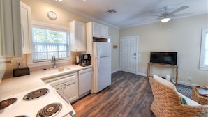 A compact kitchen with a stove, sink, and refrigerator, a dining area with a wicker sofa, and a ceiling fan. - Beautiful, private,  bright one bedroom cottage walk to Duval, Truman Waterfront, Santiago's Bodega (Key West)