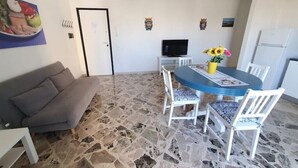 A dining area with a stone floor, a grey sofa, a round table with chairs, a TV on a stand, and a refrigerator. - Casa Blu Marino (Favignana)