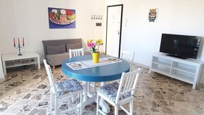 A dining area with a round blue table, white chairs, a vase with flowers, a television on a white stand, and a stone floor. - Casa Blu Marino (Favignana)