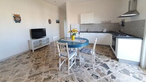 A compact kitchen with white cabinets, a built-in stove, and a dining area with a blue table and white chairs. - Casa Blu Marino (Favignana)