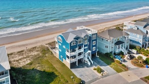 Zona residencial costera con varias casas de dos pisos, con balcones y ventanas grandes, ubicada cerca de una playa con olas visibles.