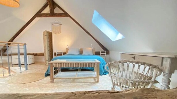 Une chambre avec un plafond en pente, un puits de lumière, un lit avec une literie bleue, un banc en bois, une chaise blanche et une petite table.