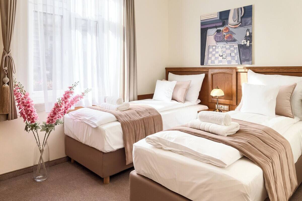 Habitación de hotel con dos camas, un ventanal con cortinas translúcidas, un cuadro en la pared y un jarrón con flores rosadas sobre el suelo.