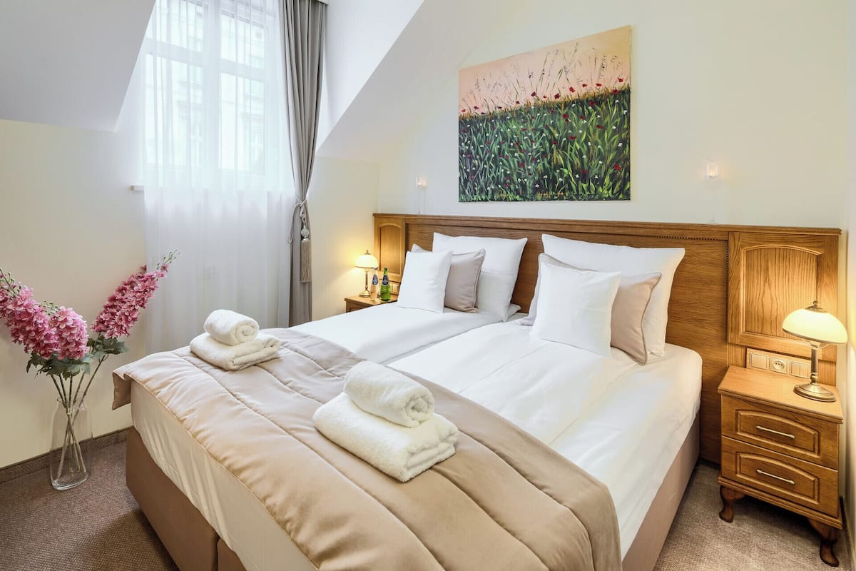 Una habitación de hotel con una cama grande, dos mesitas de noche, un florero con flores y un cuadro.