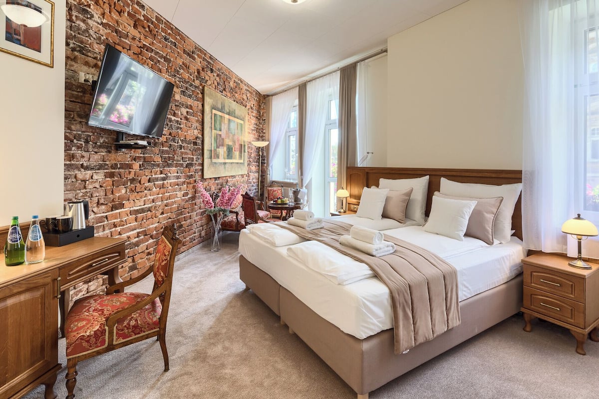 Habitación de hotel con pared de ladrillo, una cama grande con ropa blanca, un escritorio de madera con silla y un televisor montado en la pared.