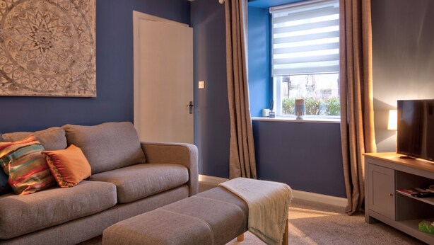 A modern living room with a sofa, ottoman, and a television on a stand. - A 2-bedroom cosy cottage based at the heart of the rural village of Brora. (Brora)
