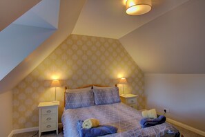 A bedroom with a sloped ceiling, two bedside lamps, a bed with a geometric patterned duvet, and a teddy bear on the bed.