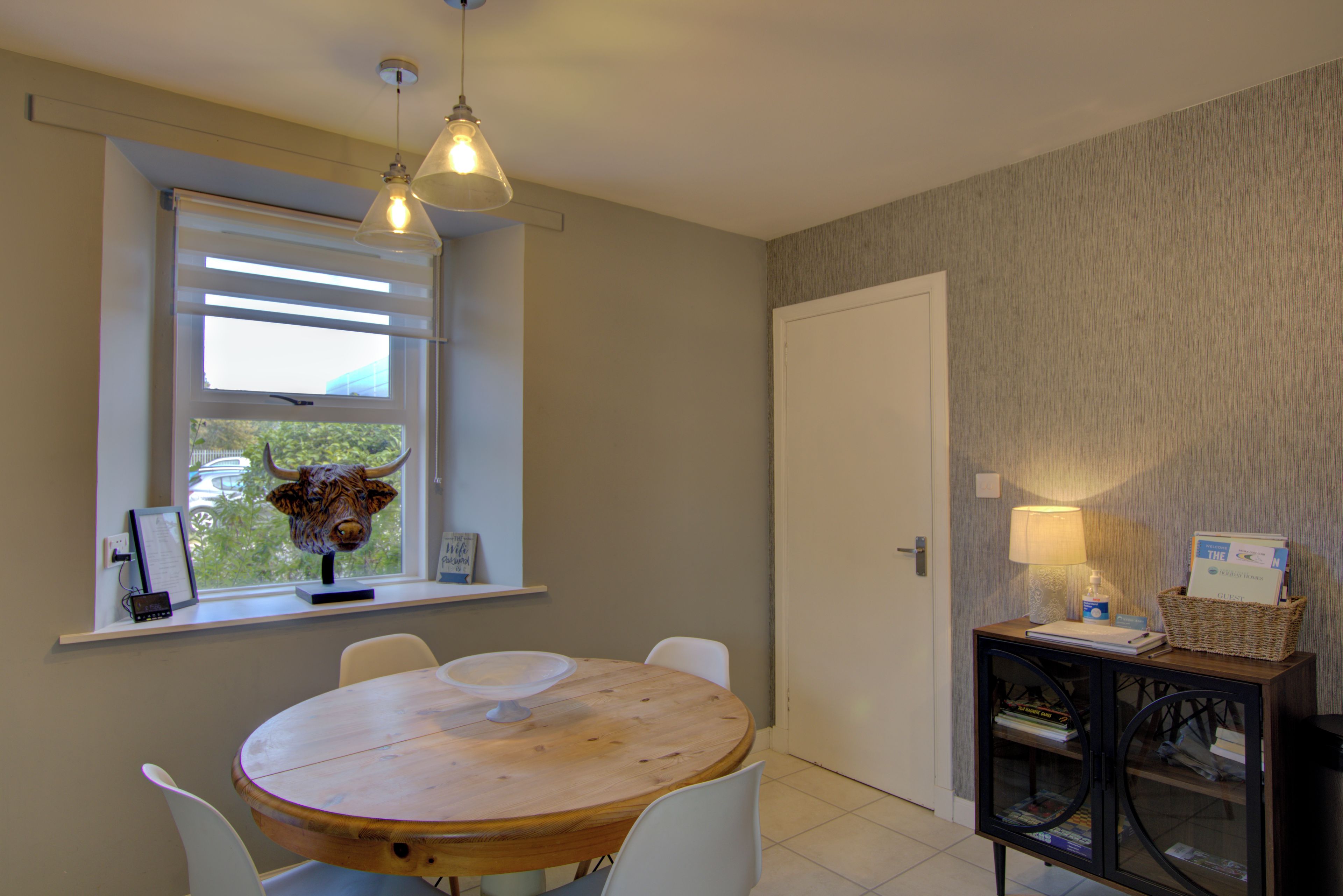 A room with a round wooden table, white chairs, a window with blinds, a wall-mounted lamp, and a small shelf with items.