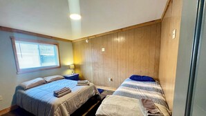 A bedroom with two beds, a window with blinds, a nightstand with a lamp, and wooden paneling on the walls.