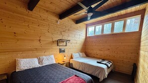 Ein blockhaushaltsähnliches Zimmer mit einem großen Bett, einem kleineren Bett, einem Deckenventilator und einem Fenster mit Blick auf Bäume.
