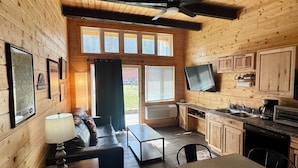 Ein Innenraum einer Hütte mit einer Couch, einem Tisch, Stühlen, einem Fernseher, einer Küchenzeile und Holzwänden.