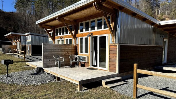 Una cabaña moderna con terraza de madera, dos sillas y un amplio ventanal corredera.