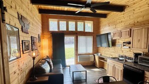 A wooden cabin interior with a sofa, dining table, kitchen, and large windows.