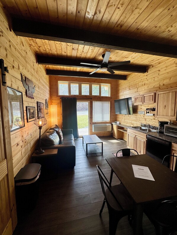 A wooden cabin interior with a sofa, dining table, kitchen, and large windows.
