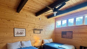 A cabin-style room with two beds, wooden walls, a ceiling fan, a window with a view, and a framed picture on the wall.