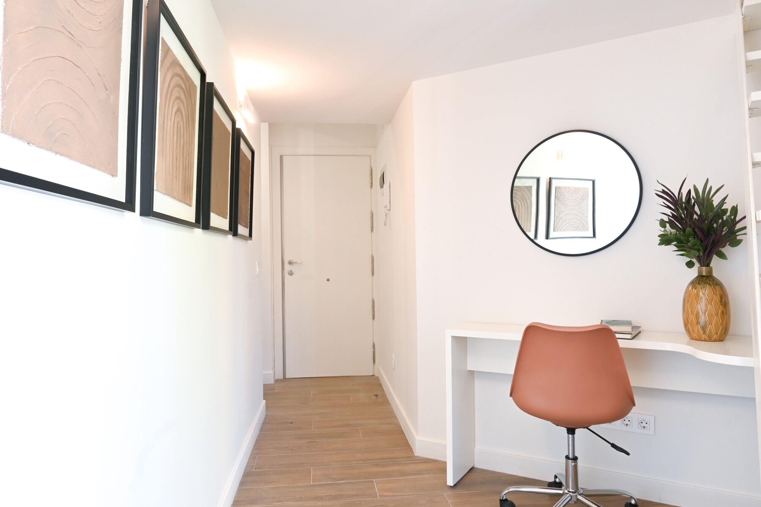 A hallway with a white wall, framed artwork, a white desk, a round mirror, and a potted plant.