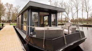 A modern floating houseboat with a covered deck, large windows, and a seating area. - Houseboat in Offingawier With Sea View (Offingawier)