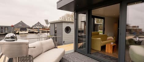 Una terraza moderna al aire libre con un sofá y una mesita, un ventanal de vidrio que da a un espacio interior con un sofá y una mesa de centro, y una vista de barcos y casas al fondo.