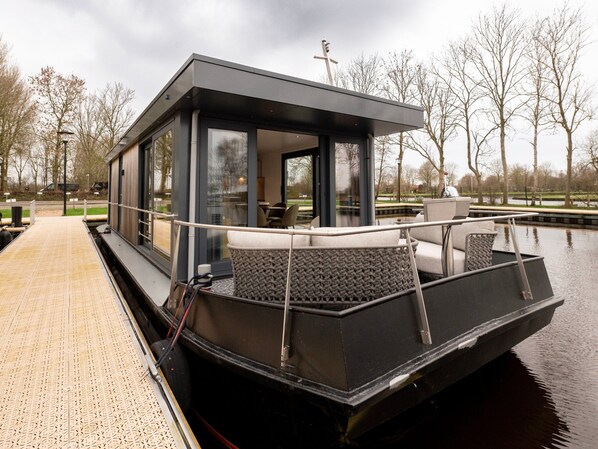 A modern floating houseboat with a covered deck, large windows, and a seating area. - Houseboat in Offingawier With Sea View (Offingawier)