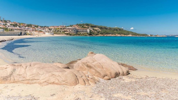 A coastal beach with clear blue waters, rocky formations, and a hillside with buildings. - Holiday Home in Baja Sardinia Near Beaches (Arzachena)