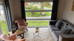 A modern living room with a grey sofa, a pink armchair, and a wooden coffee table. A large window offers a view of a green garden and a street. - Charming Chalet With Spacious Enclosed Garden (Hippolytushoef)