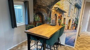 A dining area with a wooden table and green upholstered chairs, a window with black curtains, and a view of a stone building through an archway. - Charming Chalet With Spacious Enclosed Garden (Hippolytushoef)
